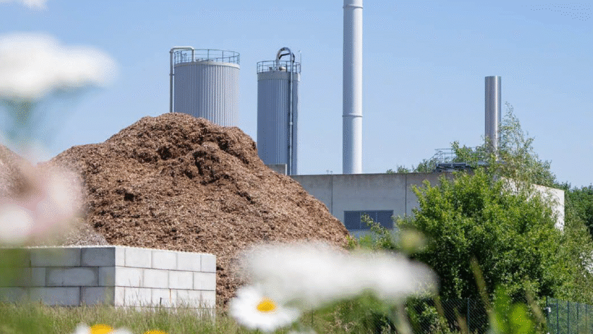 Industrieanlage mit einem hohen Schornstein und einem Holzlager im Vordergrund, umgeben von Blumen. TEAtherm in Dinkelsbühl