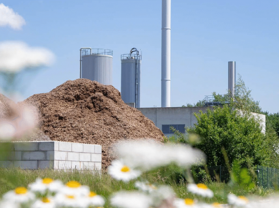 Industrieanlage mit einem hohen Schornstein und einem Holzlager im Vordergrund, umgeben von Blumen. TEAtherm in Dinkelsbühl