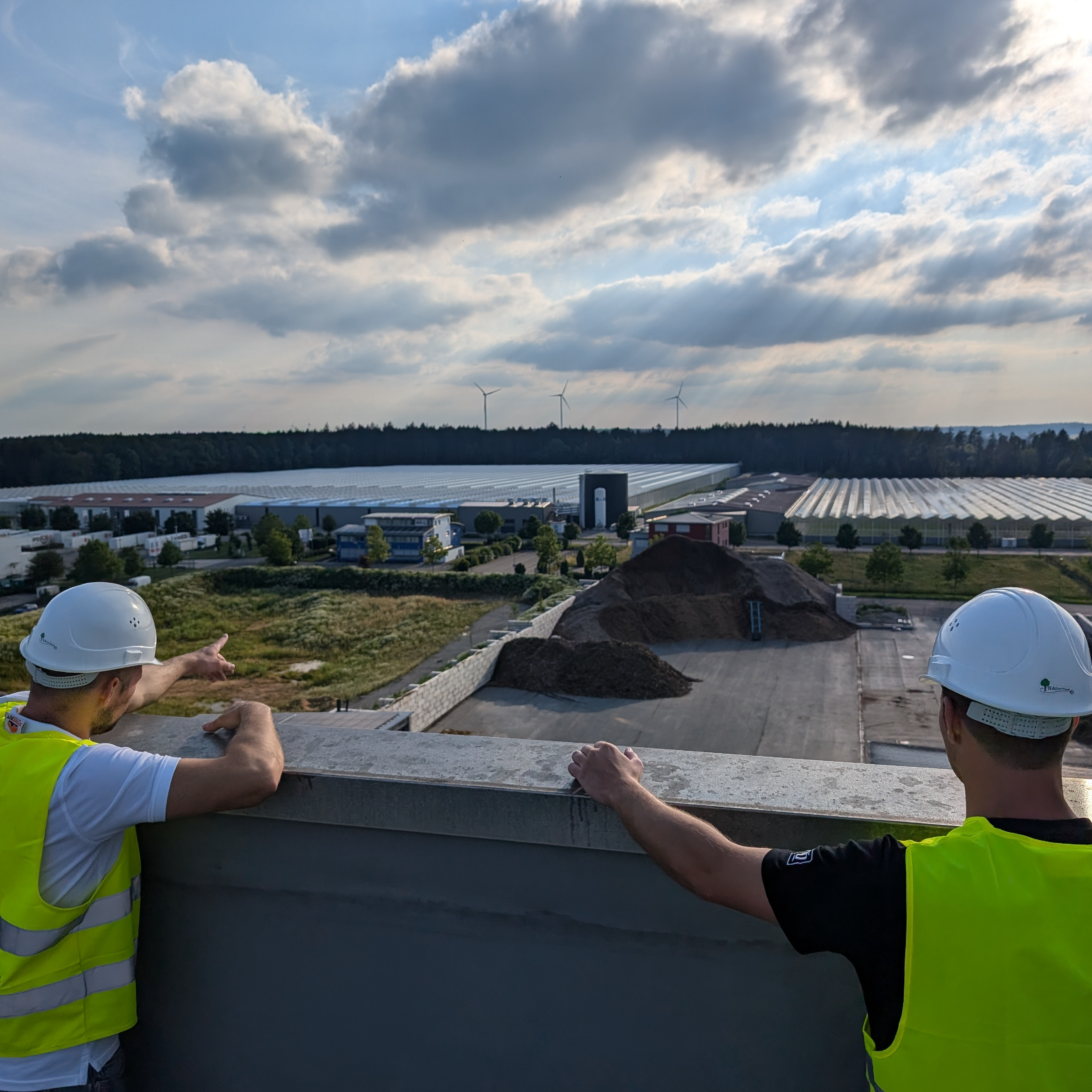 Zwei Personen in Sicherheitswesten und Helmen stehen auf einem Dach und schauen auf ein Gelände mit Windrädern im Hintergrund. TEAtherm in Dinkelsbühl
